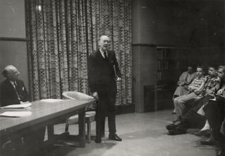 Picture of man standing at a desk and speaking to an audience. 