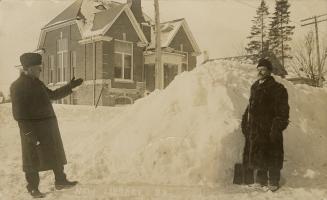 Two men standing in front a huge mound of snow. Gabled public building in the background.