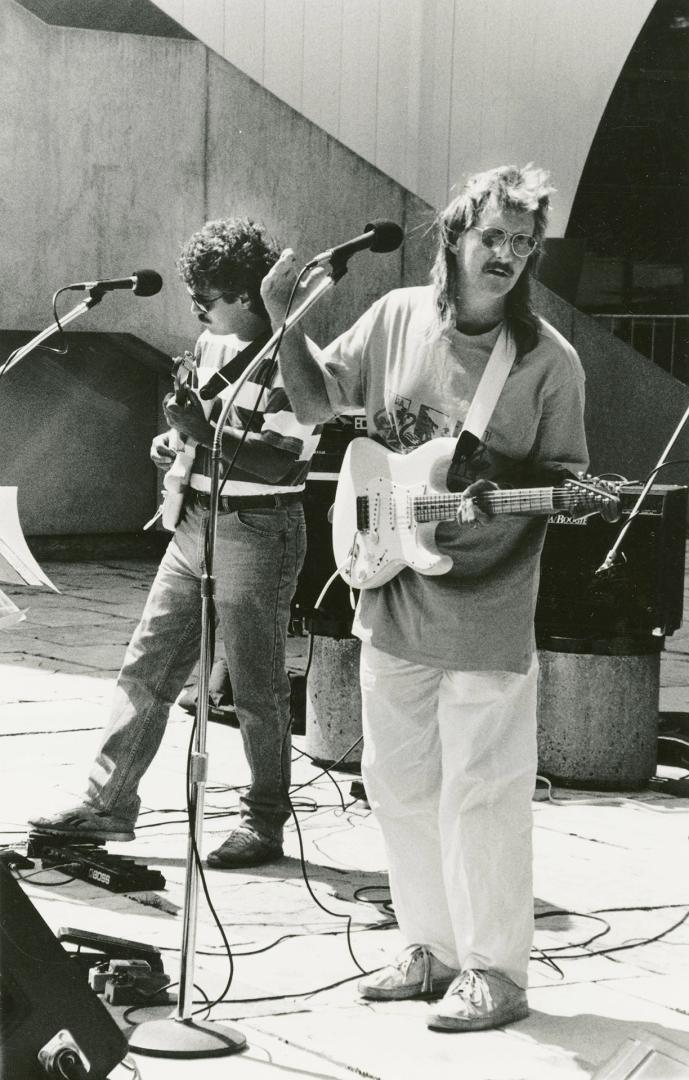 Outdoor sunshine. Two young men on guitars behind microphones in front of Brutalist concrete st…