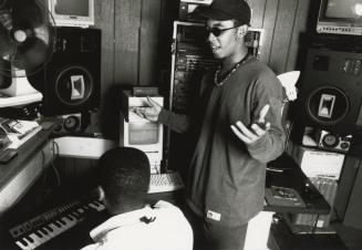 Young BIPOC man stands with hand up above another young BIPOC man on a keyboard in a small wood…