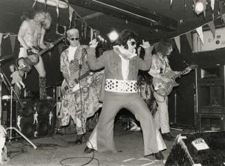 On darkened stage, four band members dressed outrageously, from left, bass player in bikini bri…