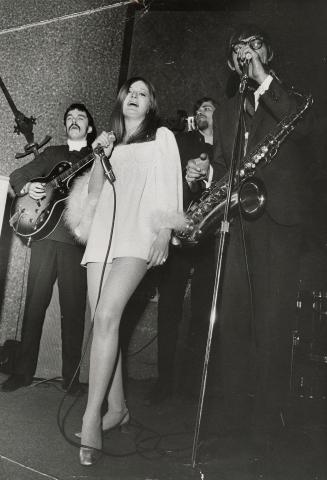 On darkened stage, young woman in white mini dress singing with microphone. Beside her, a young…