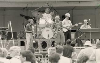 Daytime on an outdoor stage, five middle-aged musicians play from left trombone, drums, saxopho…
