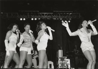 From the knees up, four young black women in satin white shorts and tops, each with a hand held…