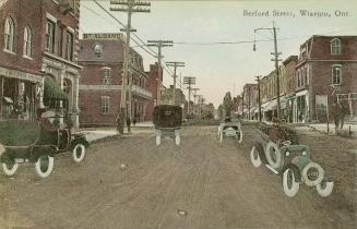 Colorized photograph of automobiles on a small town street. Buildings on either side.