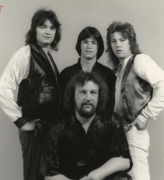 One bearded man sitting wearing black shirt. behind him three younger men with long hair. Plain…