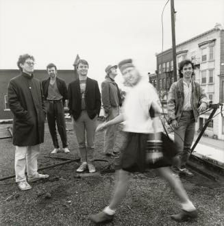 Blurry smiling young woman walks in front of five young men on a roof over a street with 4 stor…