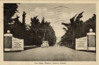 Black and white photograph of an automobile about to drive through gates spanning a roadway.
