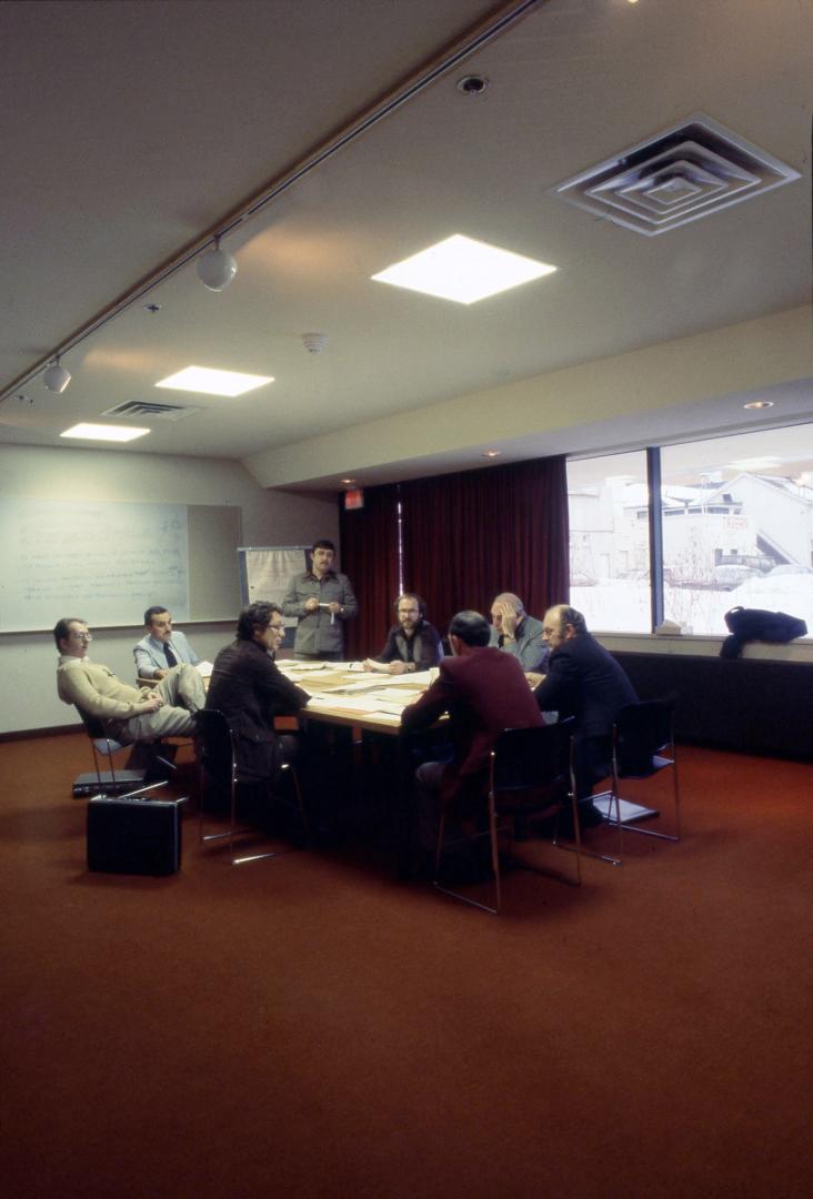 Picture of people seated around a table in a meeting room with orange carpeting and a large win…