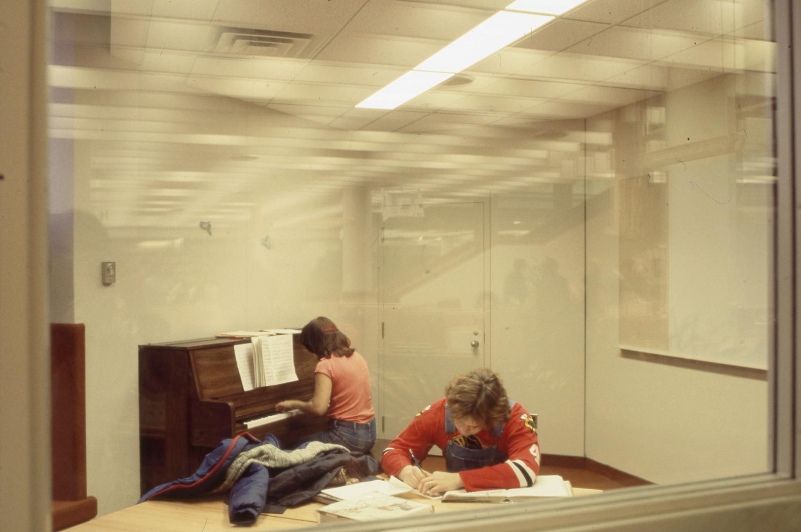 Picture of tow people in a glassed room with one playing a piano and other at a table studying.…