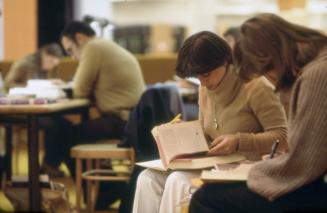 Picture of people sitting on a library bench reading and in background people at a table studyi…