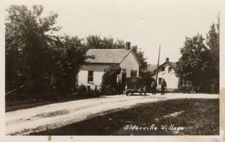 Black and white photograph of two men and an automobile in front of two small wooden buildings.