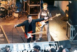 On stage, lead singer with mouth wide open and guitar, arms outstretched and another guitarist …
