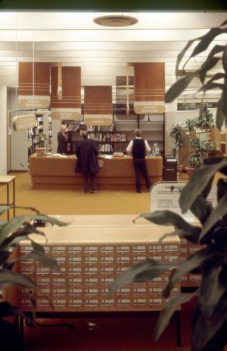 Picture of a large wooden card catalogue in foreground and reference desk with hanging signs in…