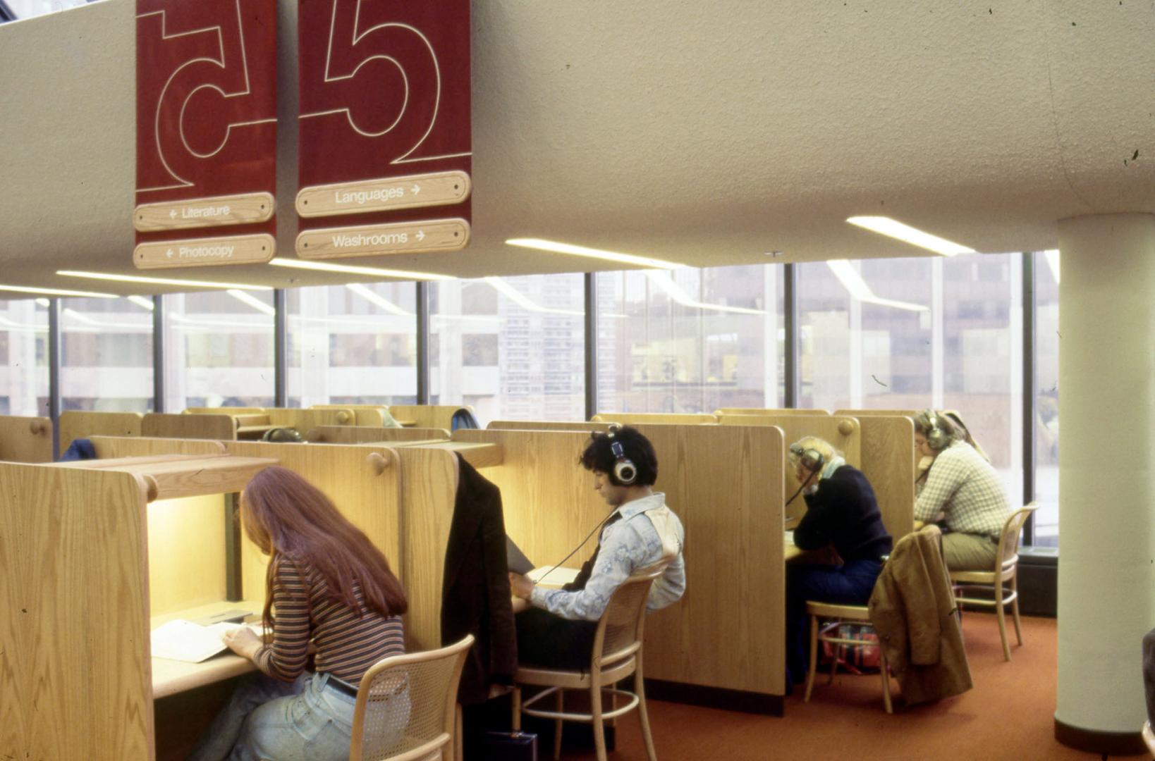 Picture of people seated at library study carrels and several are wearing headphones. 