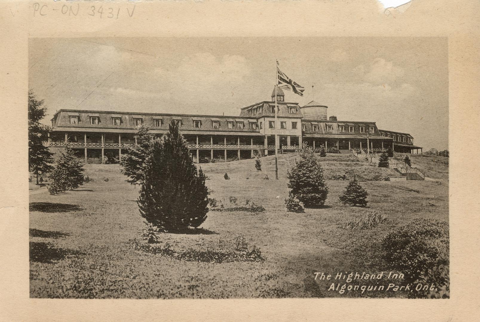 Black and white photograph of a three story hotel with a field with a flagpole in the foregroun…