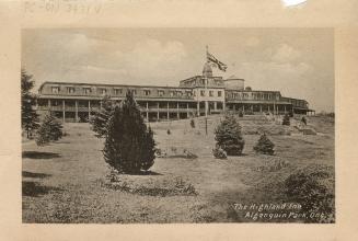 Black and white photograph of a three story hotel with a field with a flagpole in the foregroun…