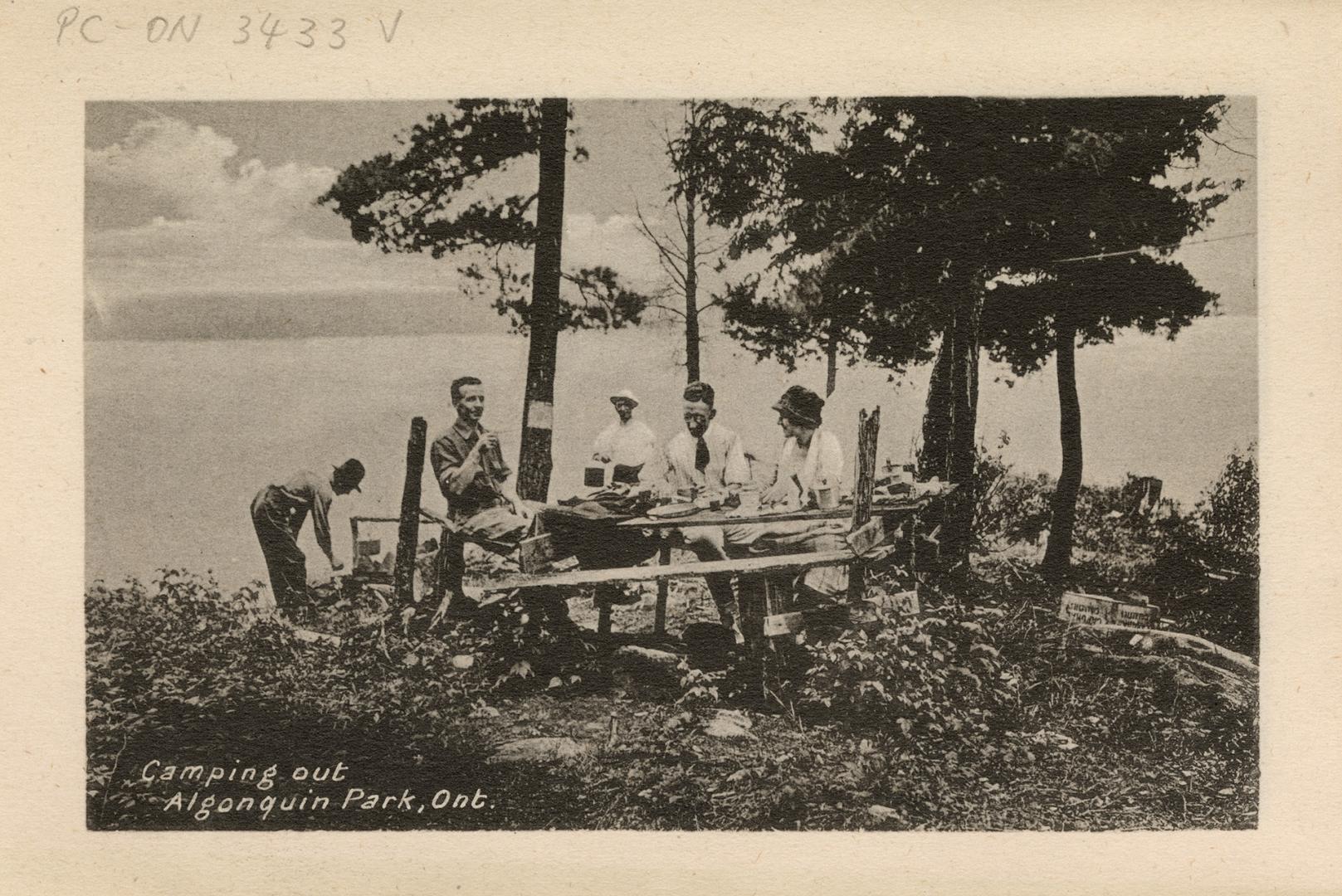 Black and white photograph of people enjoying a meal at a picnic table on rocks of a shoreline.