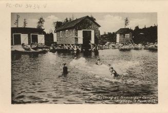 Black and white photograph of four people swimming in front of a boat house.
