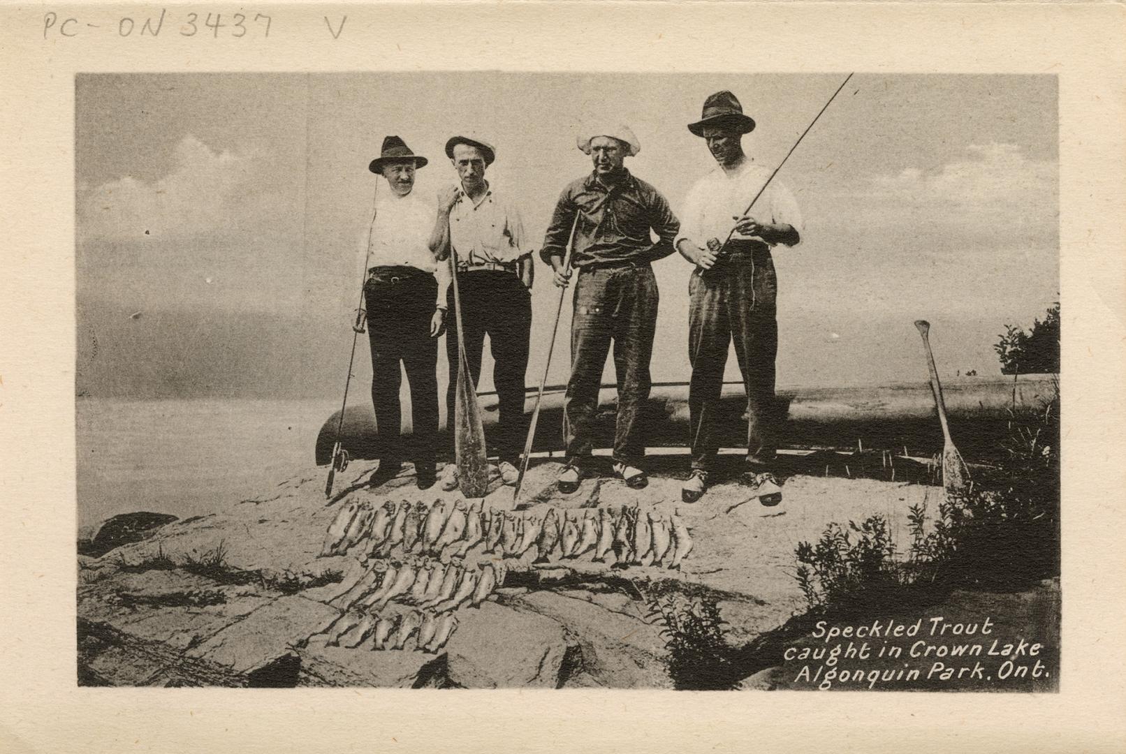 Black and white photograph of four men with canoe paddles and fishing rods standing on a rock i…