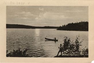 Black and white photograph of a man paddling a canoe on a lake with the sun setting behind him.