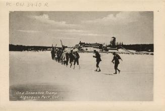Black and white photograph of a line of people snowshoeing in front of a large hotel.