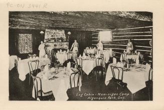 Black and white photograph of four servers standing behind formally set dining room tables in a…