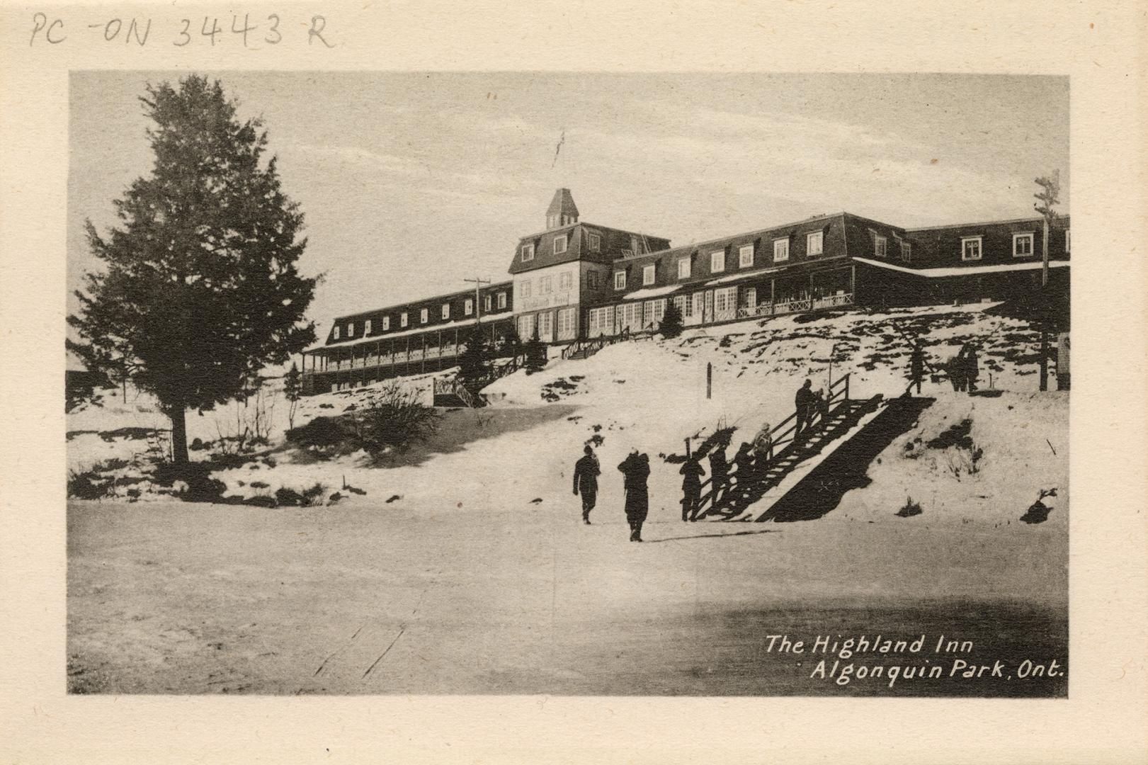 Black and white photograph of a group of people climbing up stairs towards a long, two-story ho…