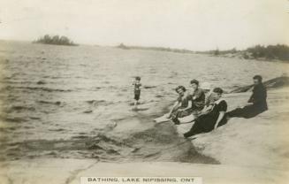Black and white photograph of a five women in bathing costumes sitting on a rock while the wave…