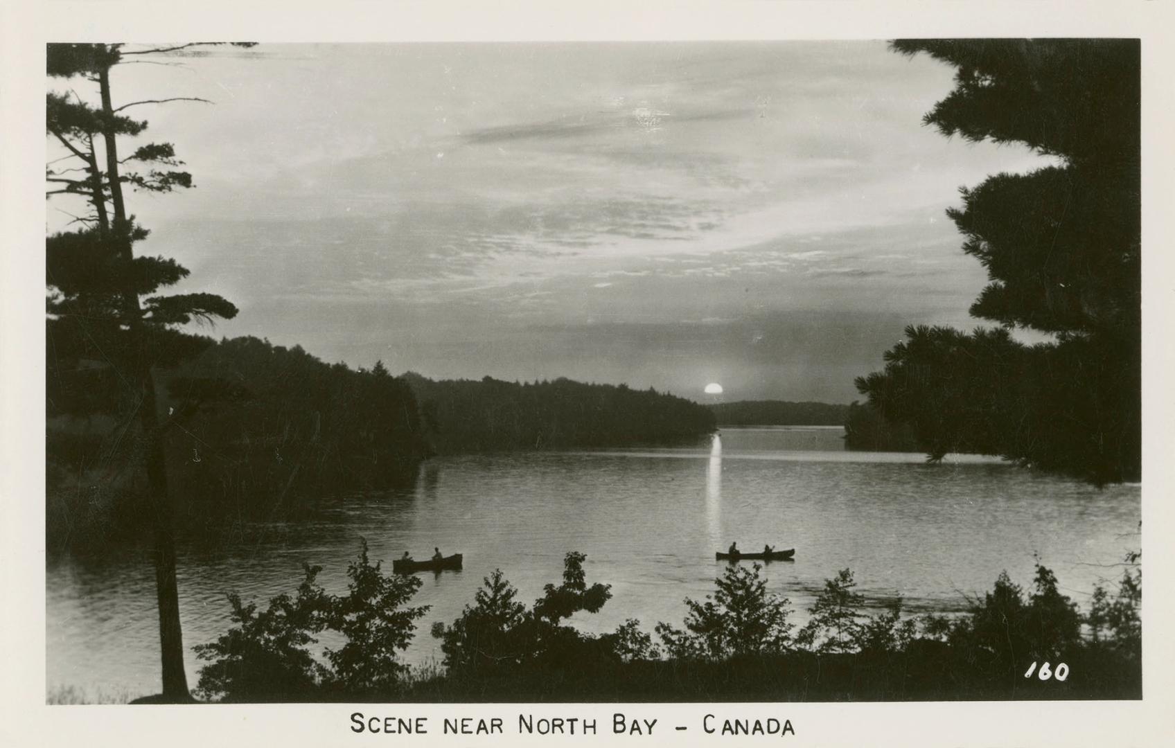 Black and white photograph of a body of water surrounded by wilderness. Three canoes in the wat…