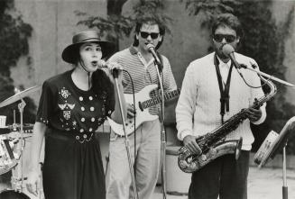 Woman in black jumpsuit and hat singing into microphone and two men to her right singing into m…