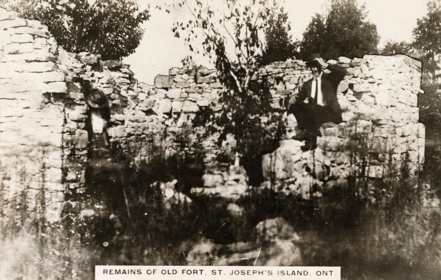 Black and white photograph of a man seated on the remains of a stone building.