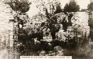 Black and white photograph of a man seated on the remains of a stone building.