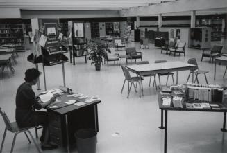 Exterior view of a library showing librarian at a desk and tables and shelves. 