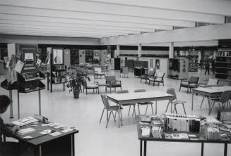 Exterior view of a library showing librarian at a desk and tables and shelves. 