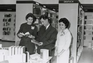 Picture of two women and a man looking at books on a table with library shelves behind them. 