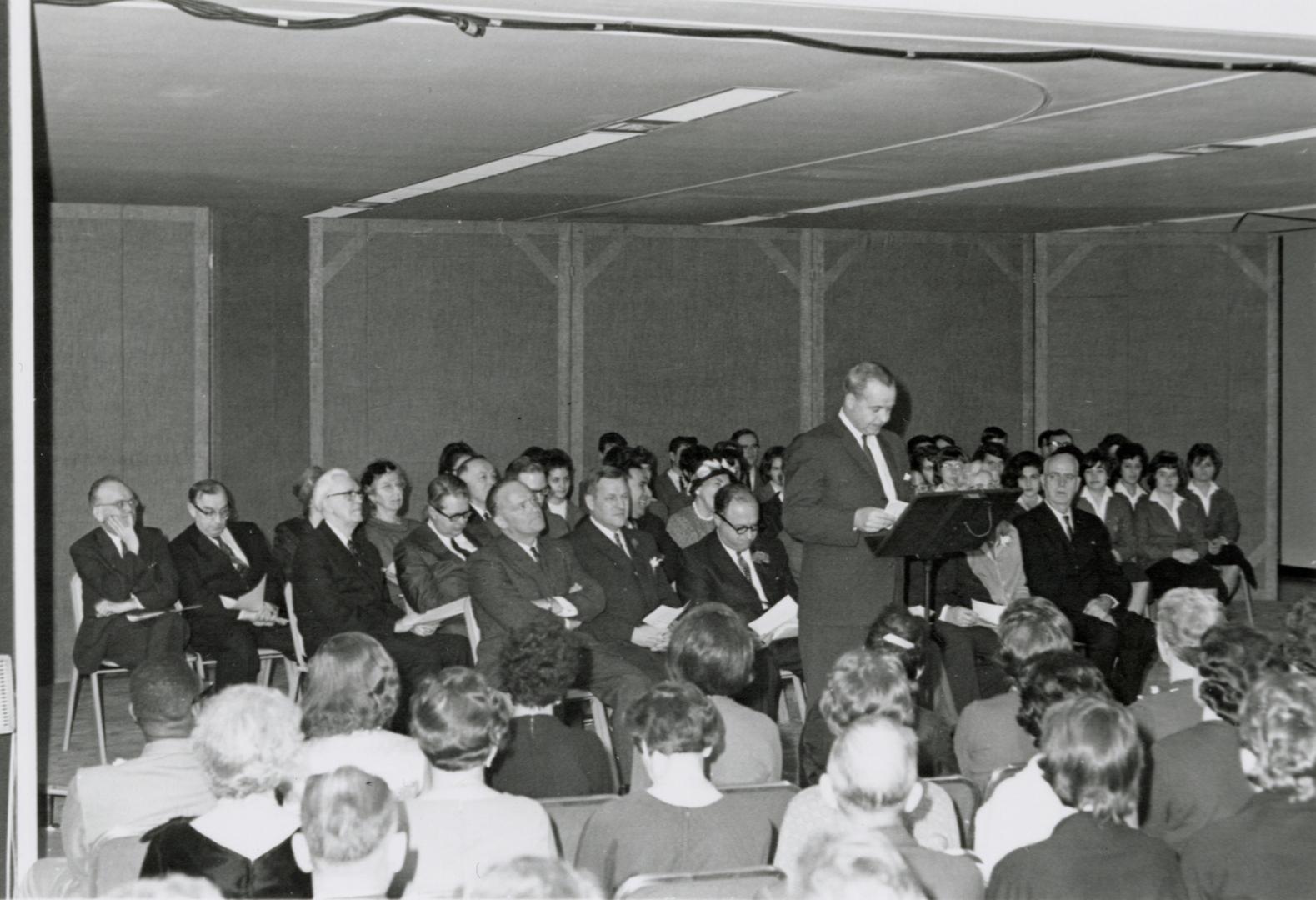 Picture of a crowd of people and man speaking at a podium. 