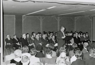 Picture of a crowd of people and man speaking at a podium. 