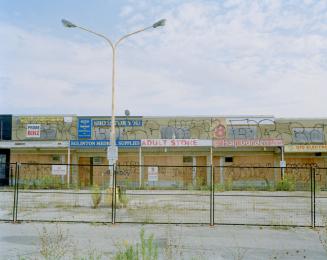 Deserted row of boarded-up storefronts covered with graffiti, behind a fence and a central ligh…