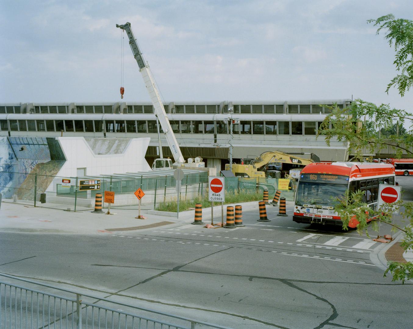 A bus drives past a construction site with a crane and barricades near Kennedy subway station.