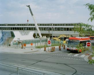 A bus drives past a construction site with a crane and barricades near Kennedy subway station.