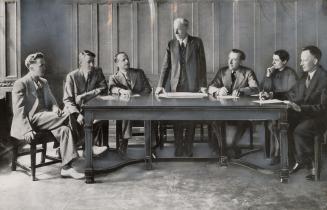 Wood paneled room with sunlight streaming in from left side window. Six men and a woman dressed…