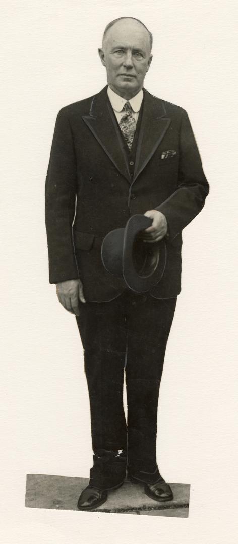 Full body, older man stands, hat in hand, wearing dark suit and white shirt and tie. 