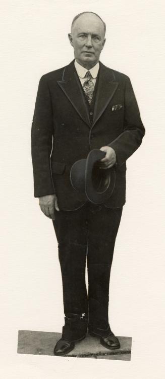Full body, older man stands, hat in hand, wearing dark suit and white shirt and tie. 