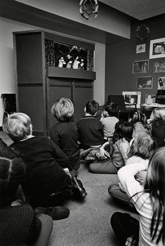 Picture of a group of children sitting on the floor watching a puppet play. 
