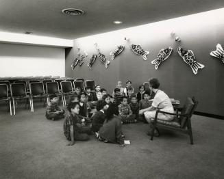 Picture of a librarian sitting in a chair telling a story to a group of children seated on the …