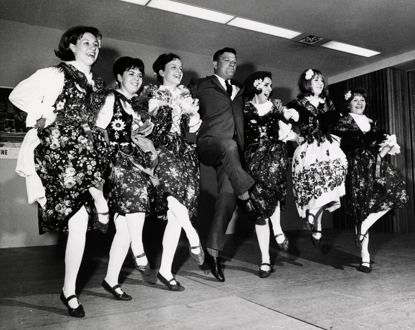 Picture of a line of six female dancers in Polish costumes with a man in the middle. 