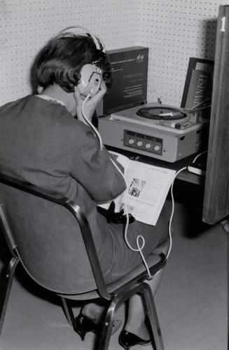 Picture of a person at a library carrel with a record player. The person is wearing headphones …