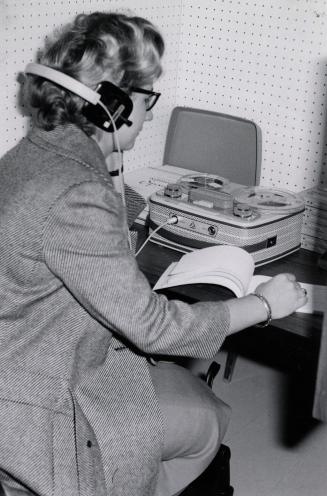 Picture of a person at a library carrel, listening to a tape deck with headphones and reading a…