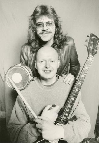 Bald young man hold a guitar and drum stick, behind him with hands on his shoulders is a young …
